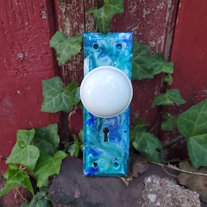 Op de afbeelding: Een blauwe en witte deurknop met een sleutelgat. De deurknop is gemaakt van hars en heeft een swirl-patroon. De deurknop is gemonteerd op een houten deur.