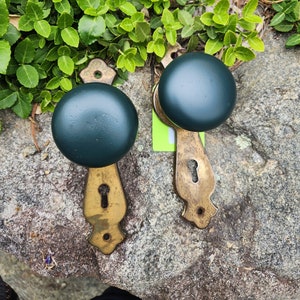 May include: Two antique brass door knobs with dark green ceramic round knobs. The knobs are on a gray rock surface.