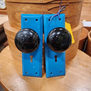 May include: Two vintage door knobs with black spherical handles and bright blue backplates. Each plate has a keyhole and three screw holes. A yellow tag is attached to one. The knobs are displayed in front of wooden boxes.