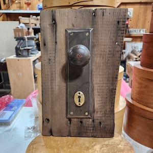 May include: A rustic wooden door plate with a vintage metal doorknob and keyhole plate. The wood has a weathered appearance, with visible grain and knots. The metal hardware has a tarnished, aged finish, adding to the antique aesthetic.