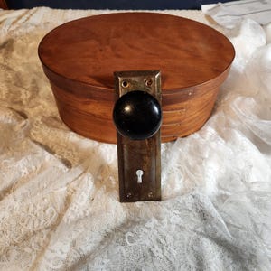 Op de afbeelding: Een antieke deurknop met een zwarte, ronde handgreep en een rechthoekige, verouderde metalen achterplaat. Op de achtergrond een ovale houten kist. De knop en achterplaat vertonen tekenen van slijtage. De achtergrond is een witte kanten stof.