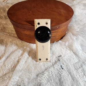 Op de afbeelding: Een antieke deurknop met een zwarte, ronde handgreep en een crèmekleurige achterplaat. De achterplaat heeft een sleutelgat en is gemonteerd op een ovale houten kist. De kist is bruin en heeft een deksel.