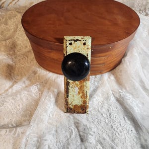 May include: An oval wooden box with a dark finish and a black knob and metal plate. The metal plate has a distressed, aged appearance with a light green paint and rust. The knob is round and black. The box is on white lace fabric.