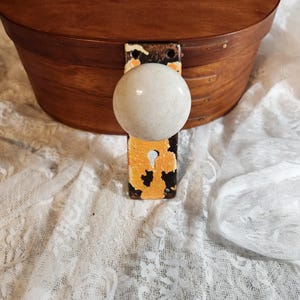 Op de afbeelding: Een ovale houten kist met een lichtgekleurde, ronde knop en een metalen scharnier. De kist is bruin met een licht verweerde afwerking. De knop is gebroken wit en het metalen scharnier heeft oranje en zwarte verf.