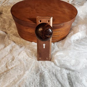 Op de afbeelding: Een antieke deurknop met een donkerbruine, patroonhouten knop en een rechthoekige koperkleurige plaat. De knop is gemonteerd op een houten kist met een rond deksel. De achtergrond is een witte kanten stof.