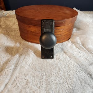 Op de afbeelding: Een ovale houten kist met een donkerbruine afwerking en zwart beslag met knop en plaat. De kist staat op witte kanten stof.