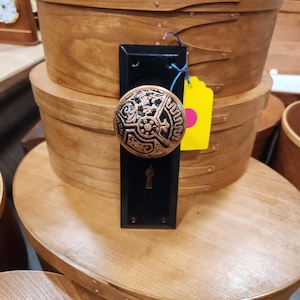 Op de afbeelding: Een zwarte deurknop met een sierlijk, koperkleurig ontwerp. De knop is gemonteerd op een zwarte rechthoekige plaat. De afbeelding toont ook verschillende houten opbergdozen met deksels, op elkaar gestapeld.