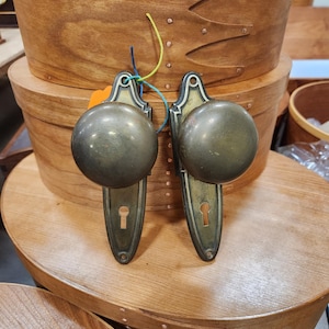 May include: Two antique brass door knobs with a round shape and keyhole detail. The knobs are attached to a decorative backplate. The knobs are displayed on a wooden surface, with wooden storage boxes in the background.
