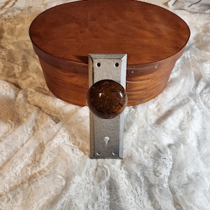 Op de afbeelding: Een ovale houten kist met een donkerbruine afwerking en een metalen deurklink met een bruine, gemarmerde knop. De klink is bevestigd aan een rechthoekige metalen plaat met een sleutelgat.