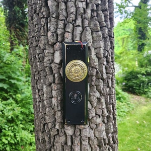 Puede incluir: Un llamador de puerta de madera negra con una perilla de metal dorado, fijado a un tronco de árbol.