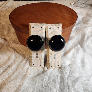 Op de afbeelding: Twee vintage deurknoppen met zwarte, ronde handgrepen en decoratieve achterplaten. De achterplaten hebben een crèmekleurige basis met een blauw-wit wervelpatroon. Een ovale houten kist staat op de achtergrond.