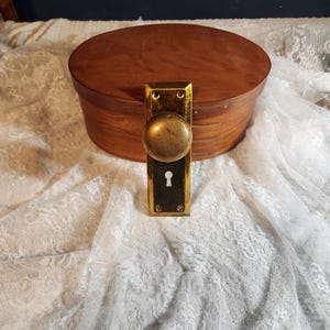 Op de afbeelding: Een vintage messing deurknop en plaat met een ronde handgreep en sleutelgat. De knop is gemonteerd op een rechthoekige plaat. Een houten ovale doos staat op de achtergrond.