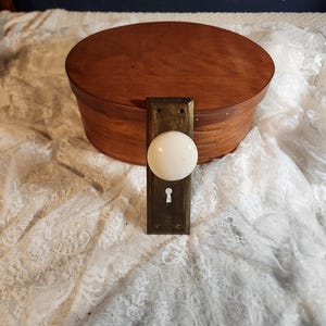 Op de afbeelding: Een vintage ovale houten kist met een messing deurklink en een witte porseleinen deurknop. De kist heeft een warme bruine kleur en de klink heeft een sleutelgat. De achtergrond is een witte kanten stof.