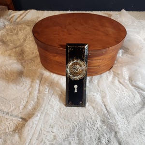 Könnte beinhalten: Eine schwarze Vintage-Türplatte mit einem dekorativen Messing-Türknauf. Der Türknauf hat ein detailliertes, kreisförmiges Design. Eine ovale Holzkiste befindet sich im Hintergrund. Die Objekte befinden sich auf einer weißen Spitzentischdecke.