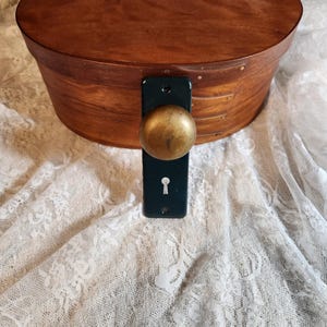 Op de afbeelding: Een ovale houten kist met een donkere afwerking en een messing knop en sleutelgatplaat. De kist staat op een witte kanten stof.