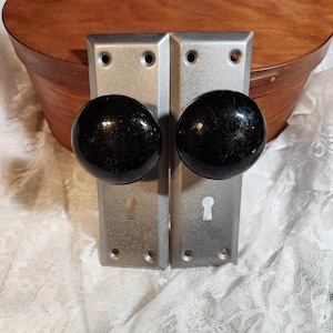 Könnte beinhalten: Zwei antike Türknöpfe mit schwarzen, kugelförmigen Griffen und silberfarbenen, rechteckigen Rückplatten. Jede Rückplatte hat ein Schlüsselloch und Schraubenlöcher. Die Knöpfe sind vor einer Holzkiste und weißer Spitze platziert.