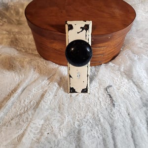 Op de afbeelding: Een antieke deurknop met een zwarte, ronde handgreep en een verweerde, gebroken witte metalen plaat. De knop is gemonteerd op een houten kist met een rond deksel. De achtergrond is een witte kanten stof.