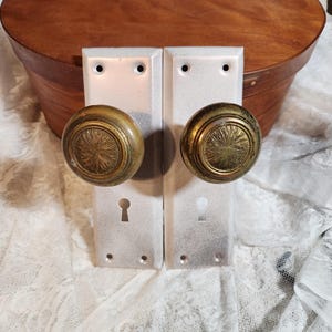 May include: Two vintage door handle sets. Each set includes a rectangular, off-white backplate with a keyhole and two screw holes. The round knobs are brass-colored with a sunburst design. A wooden box is in the background.