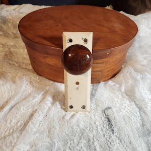 May include: An antique door knob with a cream-coloured rectangular backplate and a dark brown, round knob. The knob is mounted on a wooden box with a rounded top. The background is a white lace fabric.