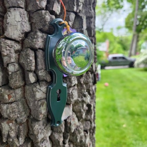 May include: A green metal door knocker with a keyhole and a round, reflective surface. The knocker is attached to a tree trunk.