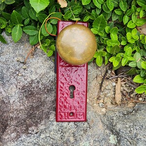 Könnte beinhalten: Ein roter Türknauf aus Metall mit einem runden Messinggriff und einem Schlüsselloch. Der Knauf ist auf einer rauen, grauen Steinoberfläche montiert.