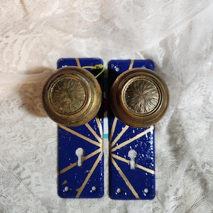 May include: Two vintage door knobs with brass hardware. The knobs are mounted on blue backplates with gold geometric designs. The background is a white lace fabric.