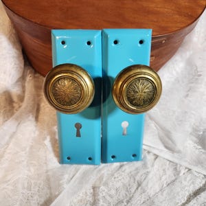 以下が含まれることがあります： 鮮やかなターコイズブルーのバックプレートと真鍮色のノブが付いた、ヴィンテージのドアノブ2つ。ノブには詳細な花の模様があります。バックプレートには鍵穴とネジ穴があります。ノブは木製の箱と白いレースを背景に配置されています。