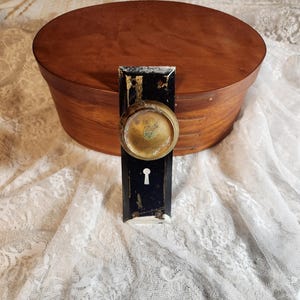 Op de afbeelding: Een vintage ovale houten kist met een donkerbruine afwerking. Een zwarte en gouden deurklink met een sleutelgat staat voor de kist. De kist en de klink staan op een wit kanten oppervlak.