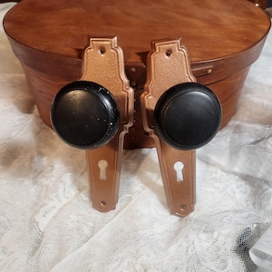 Könnte beinhalten: Zwei Vintage-Türknöpfe mit Bronze-Finish und schwarzen runden Griffen. Die Knöpfe haben ein Schlüsselloch-Design und sind vor einer Holzkiste und weißem Spitzengewebe platziert.