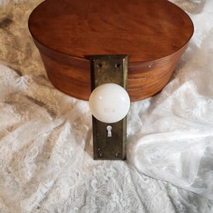 May include: A vintage wooden oval box with a brass latch and a white ceramic door knob. The box is a warm brown color, and the knob is mounted on a rectangular brass plate. The background is a white lace fabric.