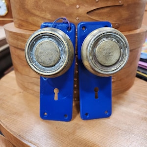 May include: Two antique door knobs with brass hardware and bright blue backplates. The knobs have a circular design and are mounted on rectangular plates with keyholes. The knobs are displayed on a wooden surface.