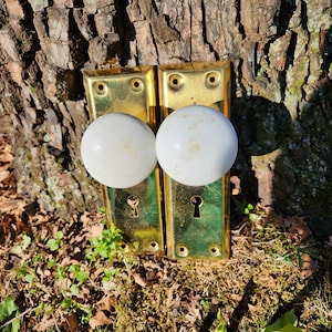 May include: Two antique brass door knobs with white porcelain handles. The knobs are mounted on a wooden surface.
