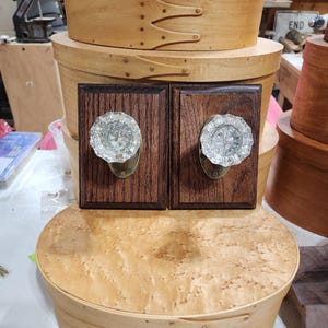 Könnte beinhalten: Zwei Holzkisten mit dekorativen Messingbeschlägen und Glasknöpfen. Die Kisten sind übereinander gestapelt, wobei die obere Kiste kleiner ist. Die Kisten sind aus hellem Holz mit einer natürlichen Oberfläche. Die Kisten stehen auf einem Tisch mit einer hellen Holzoberfläche.