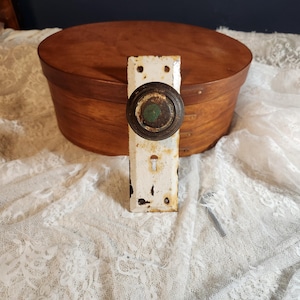 Peut inclure: Bouton de porte blanc antique avec un centre marron foncé et vert, monté sur une plaque rectangulaire. Le bouton est posé devant une boîte en bois avec un couvercle rond. Le bouton de porte montre des signes d'âge et d'usure.