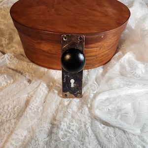 Op de afbeelding: Een ronde houten kist met een donkerbruine afwerking. Een zwarte knop en een metalen plaat met een sleutelgat zijn aan de voorkant bevestigd. De kist staat op witte, kanten stof.