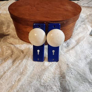 Op de afbeelding: Een ovale houten kist met een donkerbruine afwerking, gecombineerd met twee blauwe deurgrepen met witte, gespikkelde knoppen. De hardware heeft sleutelgatdetails. De items worden gepresenteerd op een wit kanten oppervlak.