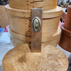 May include: A brass door plate with an oval centre design and keyhole. The rectangular plate has a tarnished finish. The background features several wooden storage boxes, suggesting a rustic or antique theme.