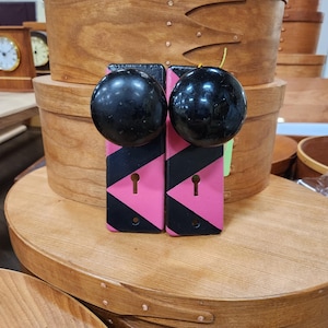 May include: Two black doorknobs with pink and black geometric accents. The doorknobs are mounted on a wooden surface, with several wooden storage boxes in the background. The boxes are oval-shaped and have a natural wood finish.