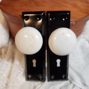 May include: Two antique door knobs with white ceramic spheres and black rectangular backplates. The knobs have keyholes and mounting holes. The background includes a wooden box and white lace fabric.
