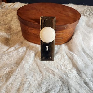 May include: An antique door knob with a white, round handle and a dark metal backplate. The backplate has a keyhole and is mounted on a wooden box. The knob is a classic design, likely from the early 20th century.
