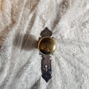 Op de afbeelding: Een vintage deurknop met een messing afwerking en een decoratieve zilverkleurige achterplaat. De knop is rond en glanzend, en de achterplaat heeft een sierlijk ontwerp. De knop staat tegen een witte kanten achtergrond.