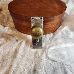 Op de afbeelding: Een ronde houten kist met een donkerbruine afwerking. De kist heeft een metalen deurklink met een messing knop. De kist staat op een witte kanten stof.