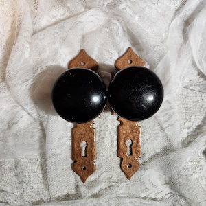 May include: Two antique door knobs with black, round handles and ornate, copper-colored backplates. The backplates have a keyhole design and a decorative shape. The knobs are set against a white lace background.