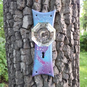 May include: A blue and purple painted door knob with a clear glass knob and a keyhole. The door knob is attached to a tree trunk.