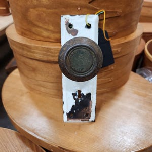 May include: An antique door knob with a distressed white backplate and a tarnished bronze knob. The knob is attached to a rectangular backplate with signs of wear and tear. A black tag hangs from the backplate. The background features wooden storage containers.