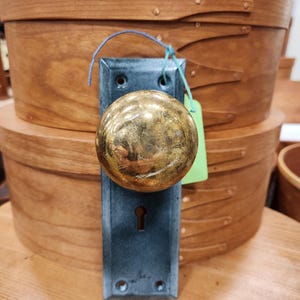 Op de afbeelding: Een vintage messing deurknop met een donkerblauwe achterplaat. De knop is rond en heeft een versleten, antieke uitstraling. De deurknop wordt getoond voor twee houten opbergdozen.