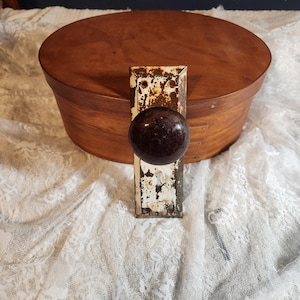 Op de afbeelding: Een antieke deurknop met een donkerbruine, ronde handgreep en een rechthoekige, roestige metalen plaat. Op de achtergrond staat een houten, ovaalvormige doos. De knop is waarschijnlijk van een vintage deur.
