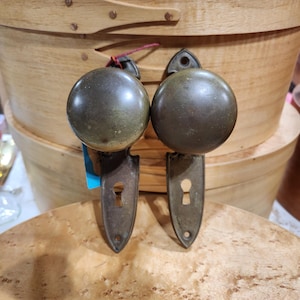 Może przedstawiać: Dwa antyczne mosiężne gałki drzwiowe z otworami na klucze i zaokrąglonym wzorem. Gałki są zamontowane na drewnianej powierzchni.