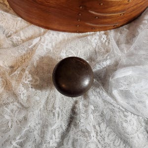 May include: A dark brown, round knob with a textured surface. The knob is set against a backdrop of white lace and a portion of a wooden box with decorative accents.