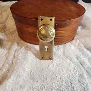 Op de afbeelding: Een vintage ovale houten kist met een messing deurknop en plaat. De kist heeft een warme bruine kleur en heeft een deksel. De messing knop en plaat zijn rechthoekig met een sleutelgat. De achtergrond is een witte kanten stof.